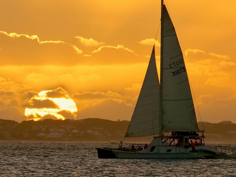 A sailing catamaran under full sail against an orange sunset
