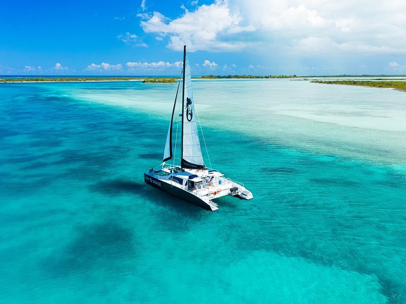 A sailing catamaran over pale turquoise flats off a white beach, seen from the air