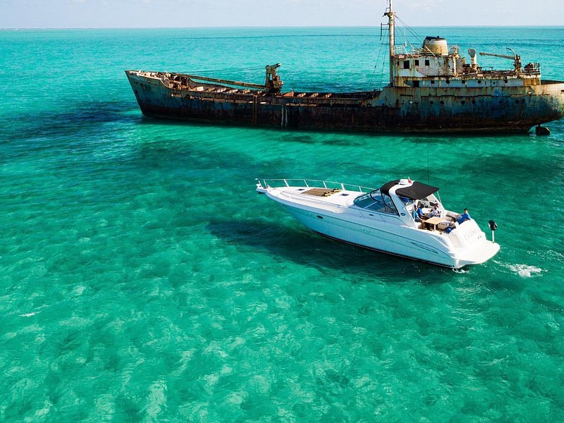 A speedboat anchored in shallow turquoise water beside a rusting shipwreck